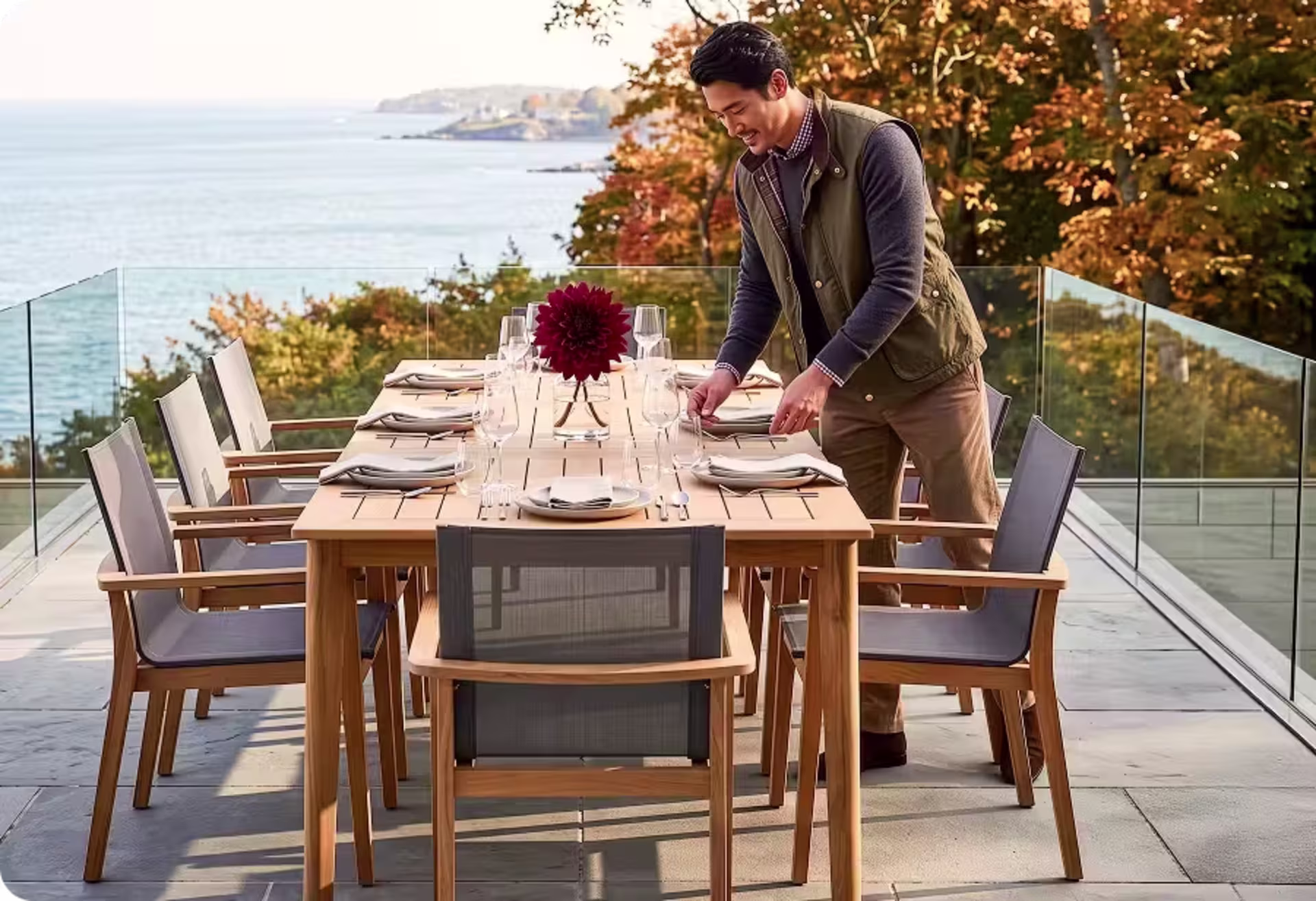 Man setting a teak outdoor dining table for a meal on a coastal patio surrounded by autumn foliage. image