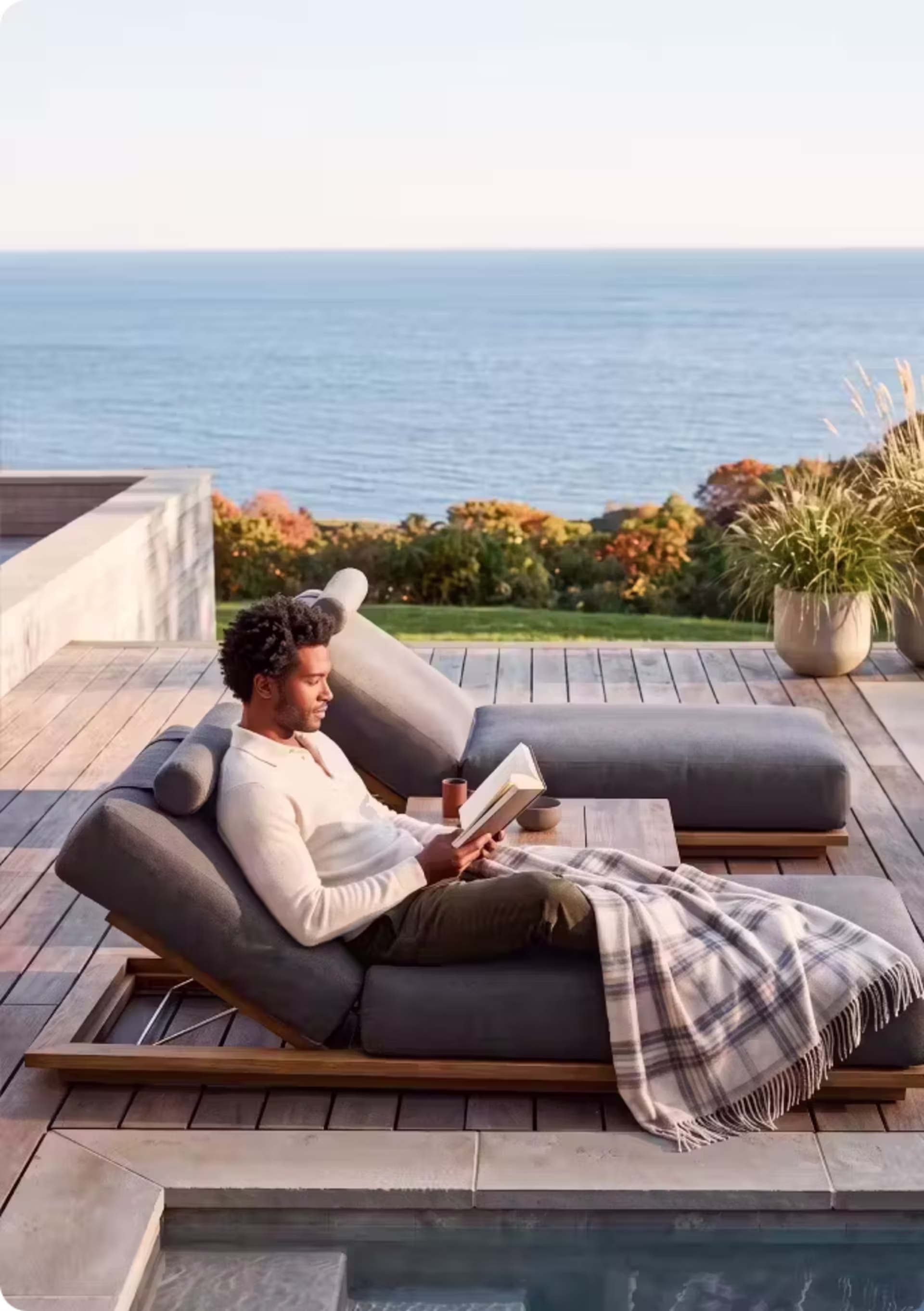 Man reading on a cushioned chaise lounge with a plaid blanket beside a pool overlooking the coast.