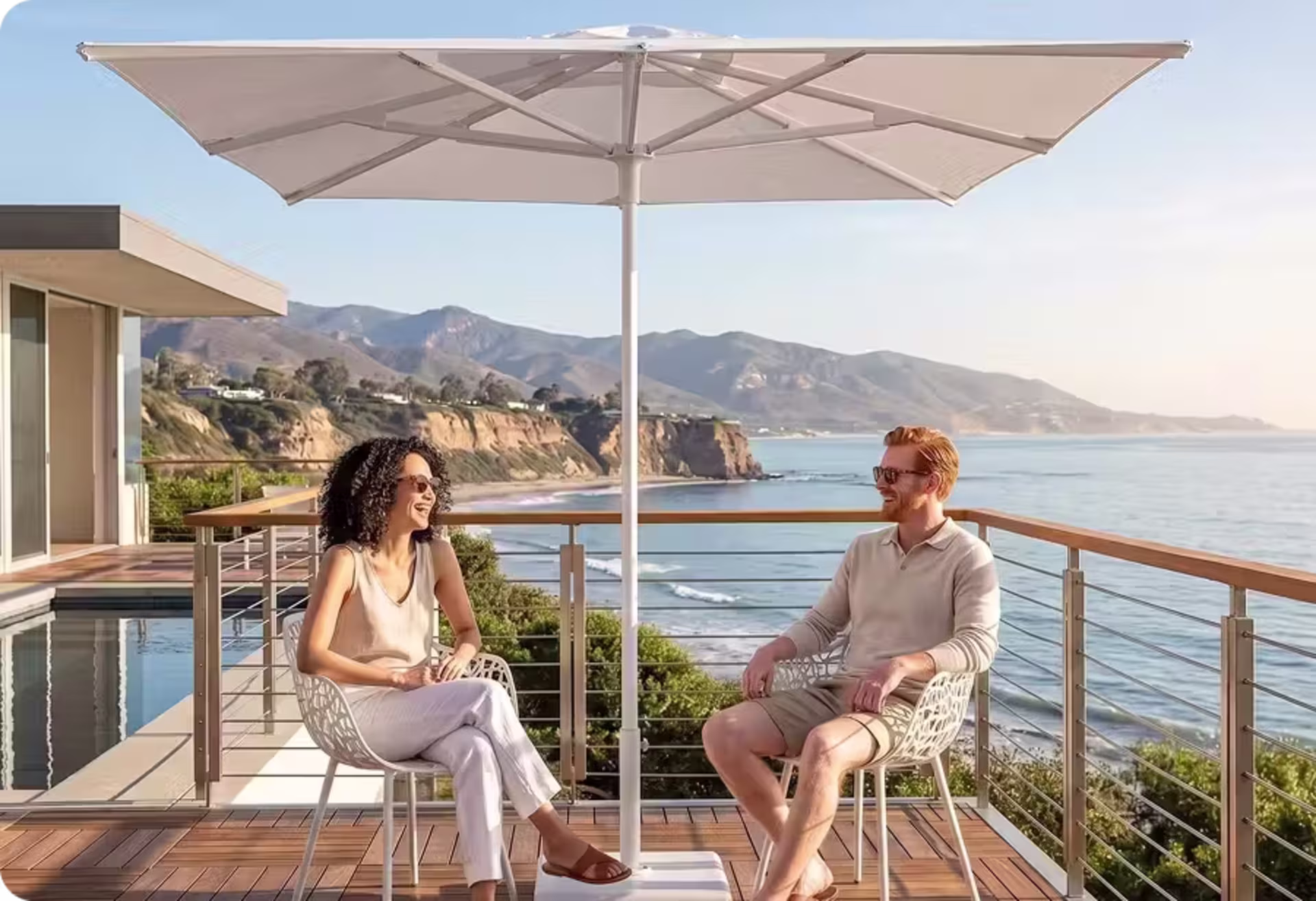 Two adults relaxing beneath a large market umbrella on a contemporary oceanfront patio with mountain and coastal views. image
