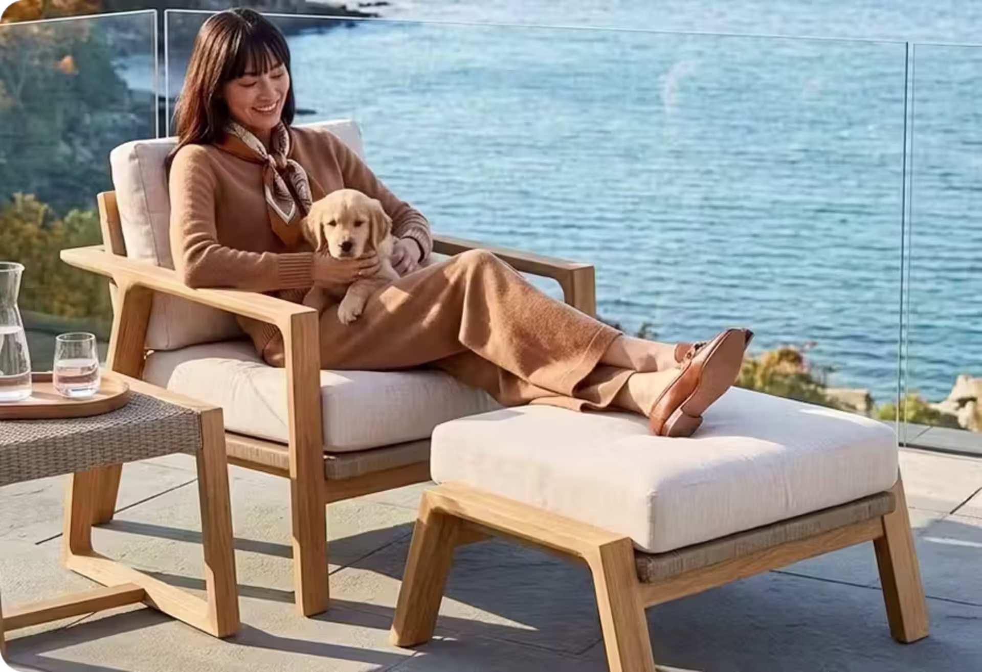 Woman relaxing with a puppy in a cushioned teak lounge chair and ottoman overlooking the coast. image
