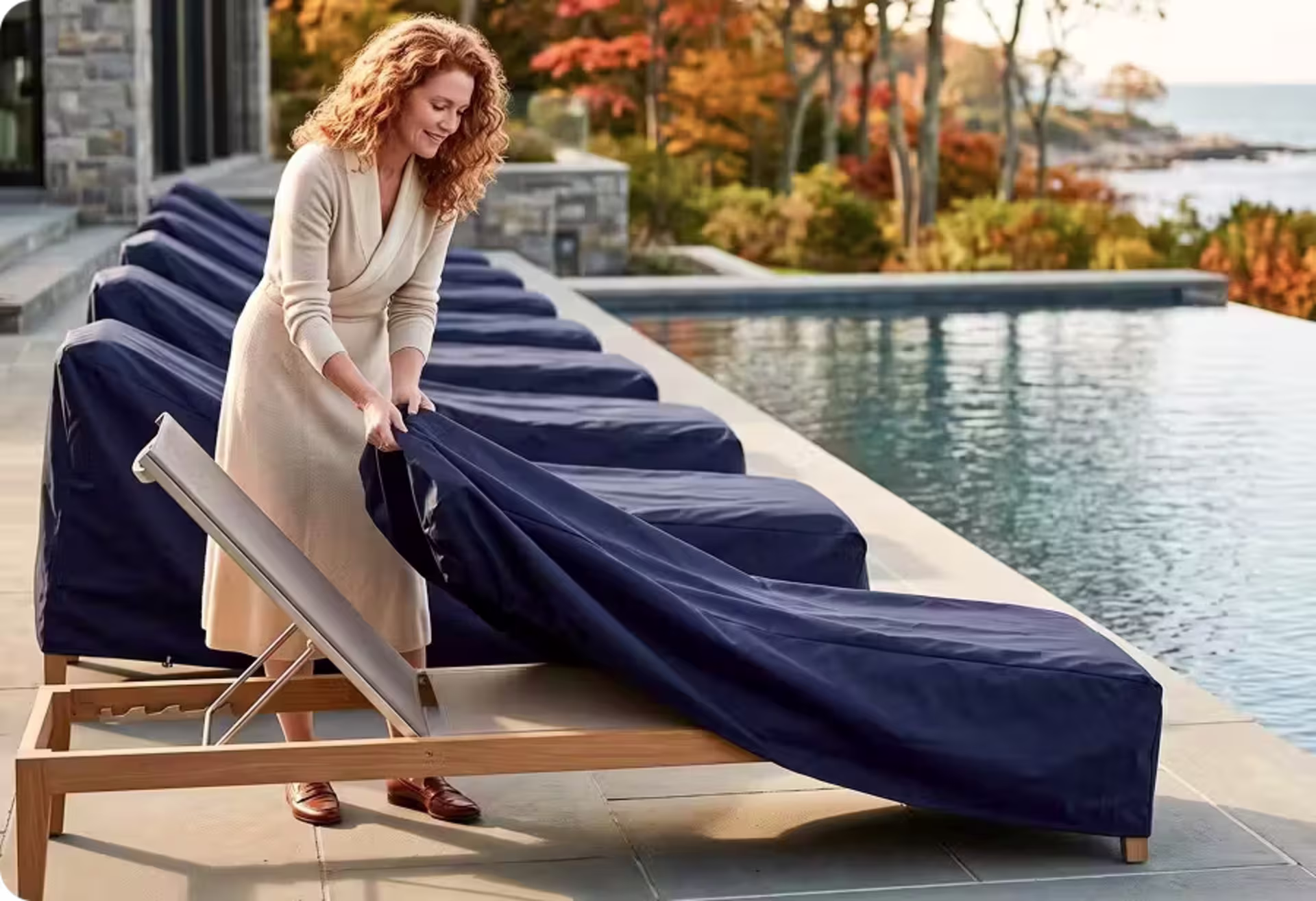 Woman covering a teak chaise lounge beside a pool with a protective outdoor furniture cover. image