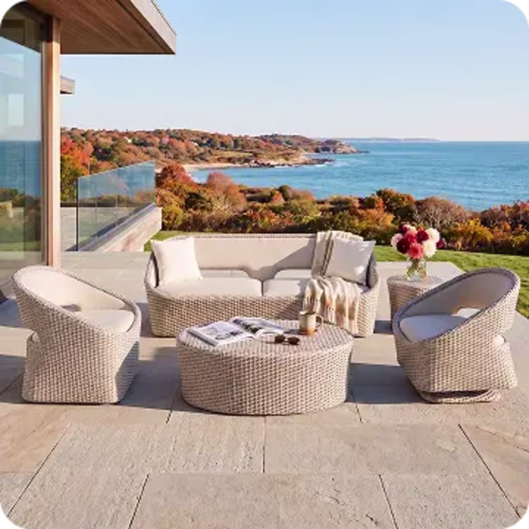Woven outdoor sofa and lounge chairs with cream cushions overlooking a coastline with autumn foliage.