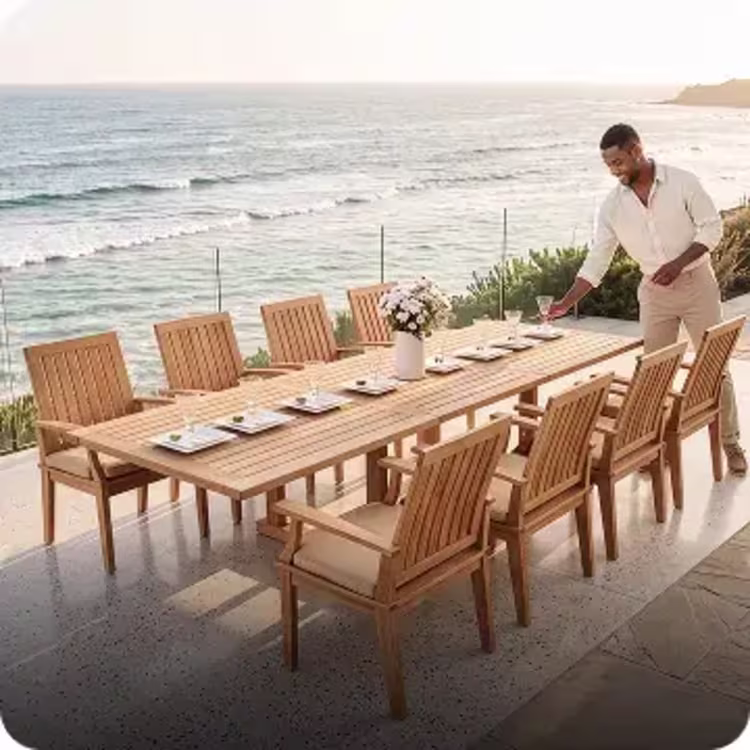 Man setting a large teak outdoor dining table overlooking the ocean.
