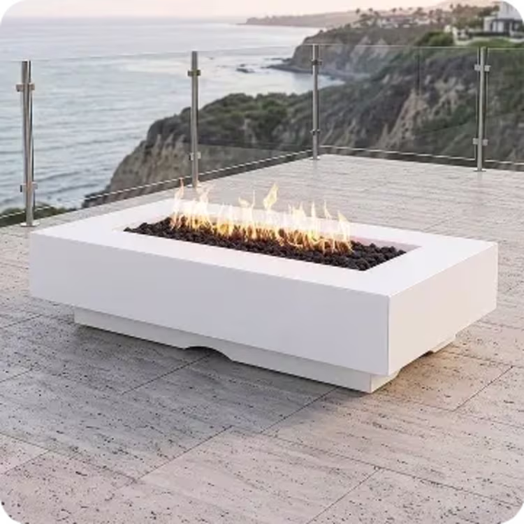 A long, low-profile rectangular white fire pit filled with black lava rock produces bright orange flames on a stone patio overlooking a coastal ocean view.