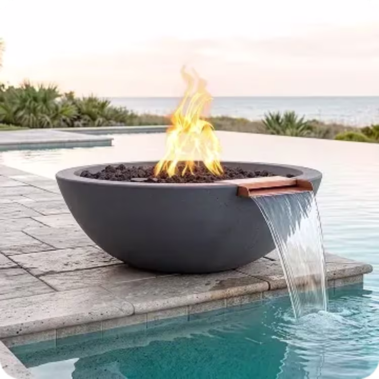 A dark grey, bowl-shaped combination fire and water feature sits on the edge of an infinity pool, with a copper spout pouring a steady sheet of water into the pool while flames rise from black lava rock in the center.