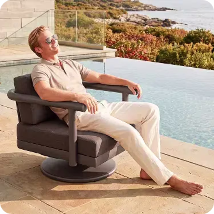Man relaxing in a cushioned swivel lounge chair beside an infinity pool overlooking the coast.