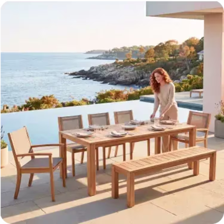 Woman setting a teak outdoor dining table for eight on a coastal patio overlooking the ocean.