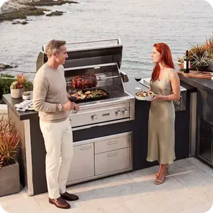 Couple grilling and serving dinner at a stainless steel outdoor kitchen overlooking the water.