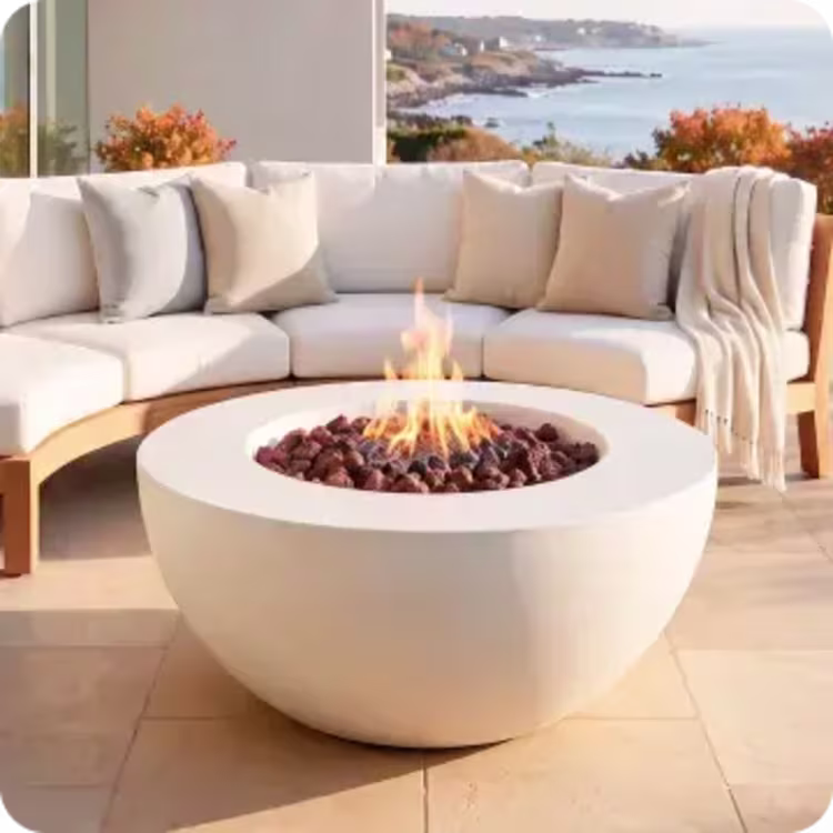 Round fire pit with glowing flames in front of a curved outdoor sectional overlooking the coast.