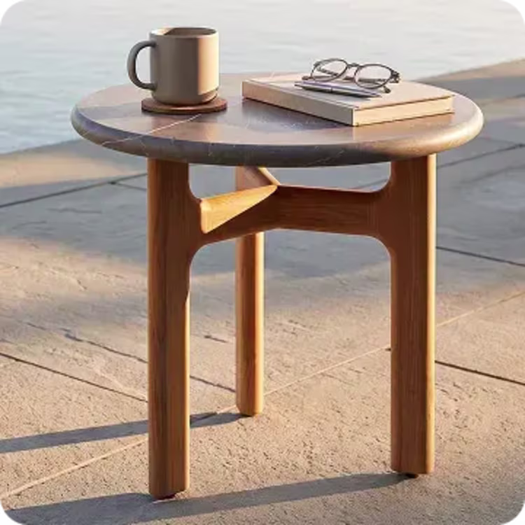 Round teak side table with a mug, book, and glasses on a sunny outdoor patio.