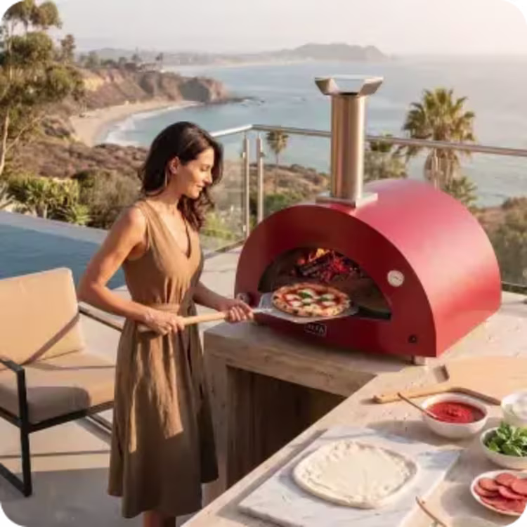 Image of a woman cooking pizza in a red outdoor pizza oven on a luxurious coastal patio overlooking the ocean.