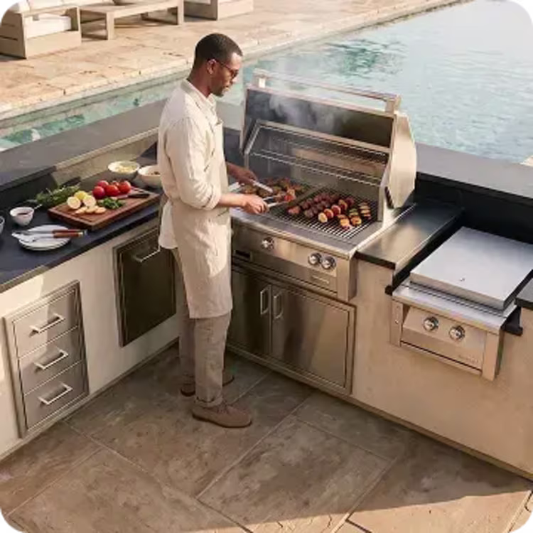 Image of a man grilling vegetables and skewers at a stainless steel outdoor kitchen beside a swimming pool.