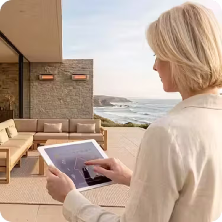 Woman uses a tablet to control wall-mounted patio heaters above a coastal outdoor lounge.