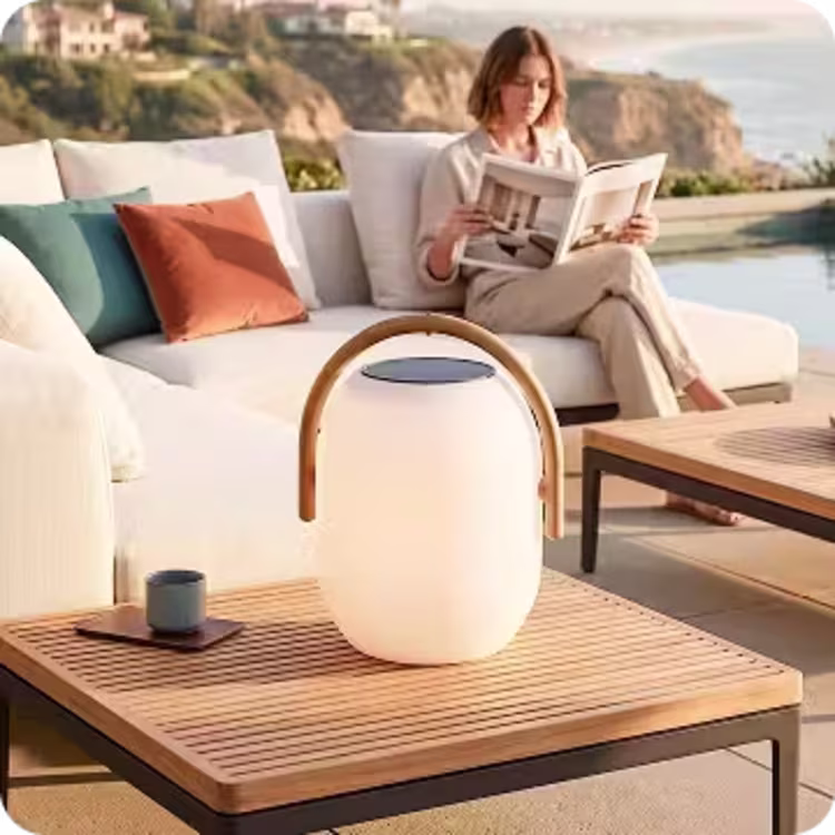 Woman reading on an outdoor sectional with a solar LED lantern on a teak coffee table overlooking the ocean.