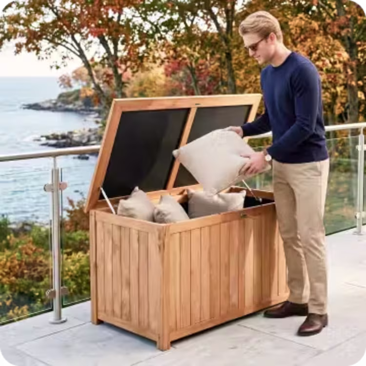 Man placing pillows into a teak outdoor storage box on a coastal patio.