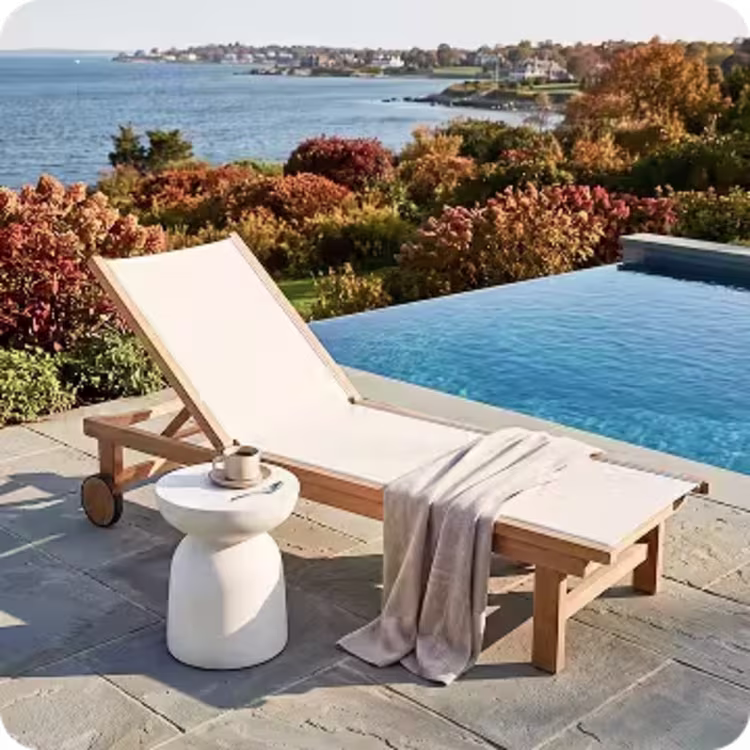 Sling and teak chaise lounge with a throw blanket and side table beside an infinity pool overlooking the autumn coast.