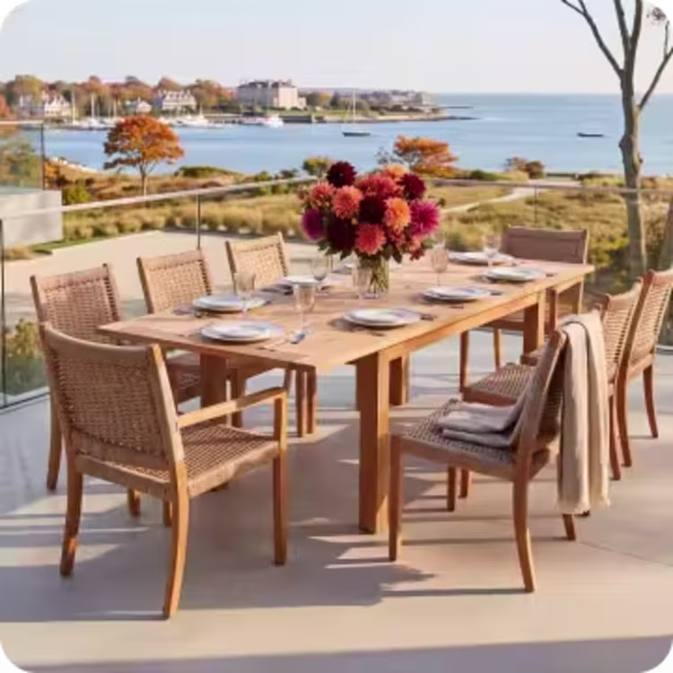 Outdoor dining table and woven chairs set for a meal with autumn flowers and a coastal view.