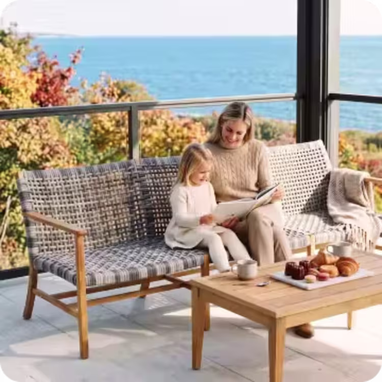 Woman and child reading together on a woven outdoor sofa beside a teak coffee table overlooking the coast.