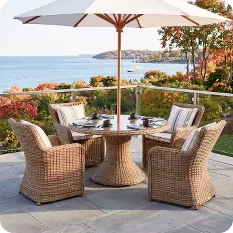 Woven outdoor dining chairs surround a round table beneath an umbrella on an autumn coastal patio.