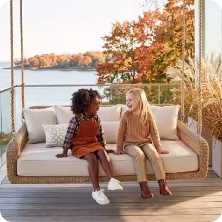 Two children sit together on a cushioned outdoor porch swing overlooking the autumn coast.