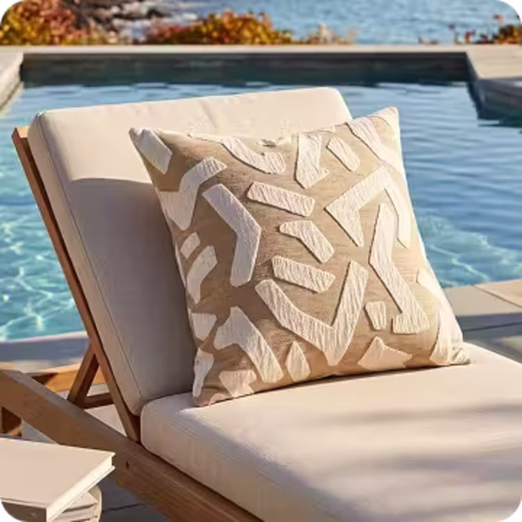 Patterned neutral throw pillow on a cushioned teak chaise lounge beside a pool.