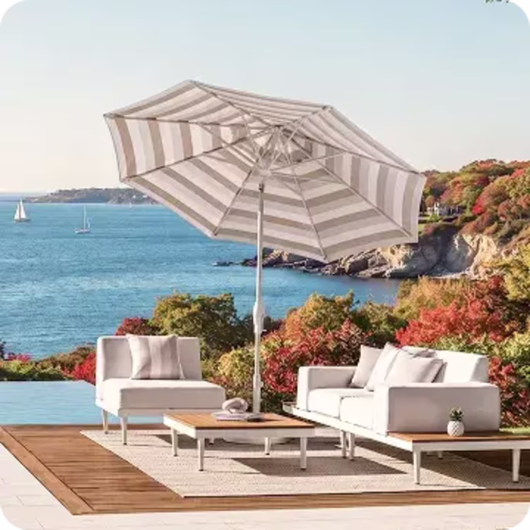 Striped patio umbrella shades a cushioned outdoor lounge set overlooking the ocean and autumn coastline.