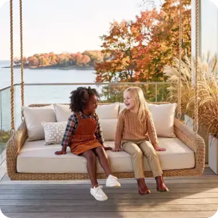 Two children sit together on a cushioned outdoor porch swing overlooking autumn foliage and the water.