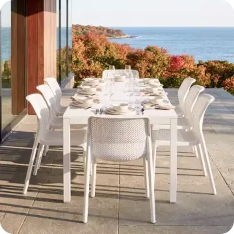 White outdoor dining table and chairs set for a meal on a patio overlooking the ocean.