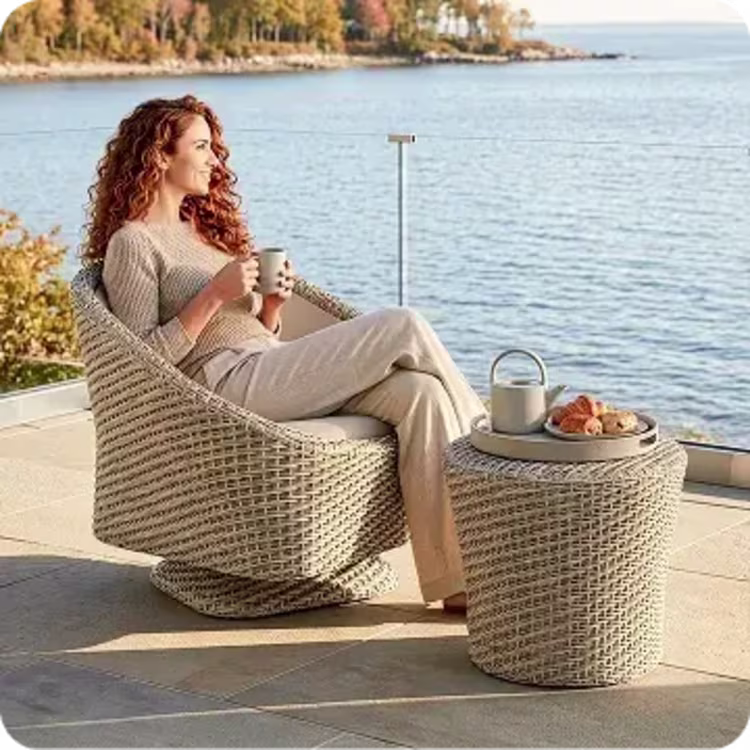 Woman relaxing in a woven lounge chair with a drink beside a coastal view.
