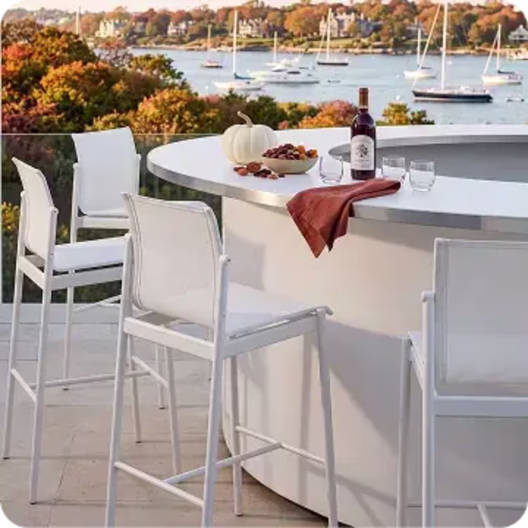 White bar stools around an outdoor bar overlooking a harbor with fall foliage.