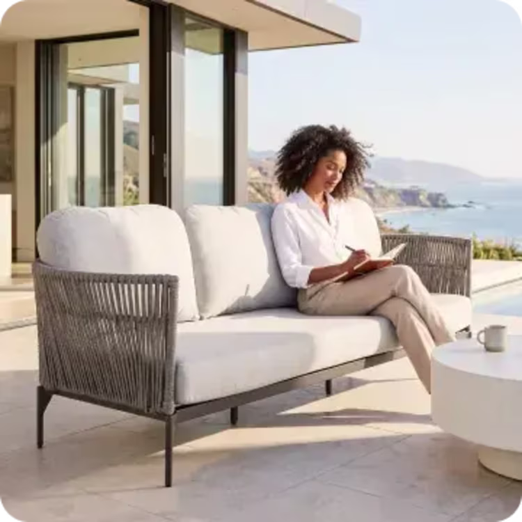 Woman reading on a woven outdoor sofa overlooking the coast.