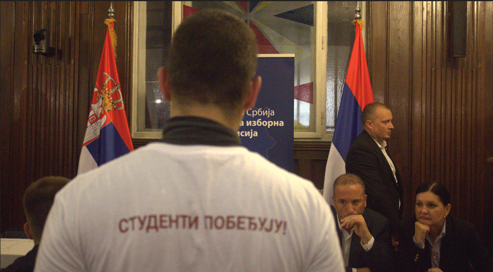 The back of a student wearing a “Students Win” T-shirt in the foreground, facing members of the Republic Electoral Commission