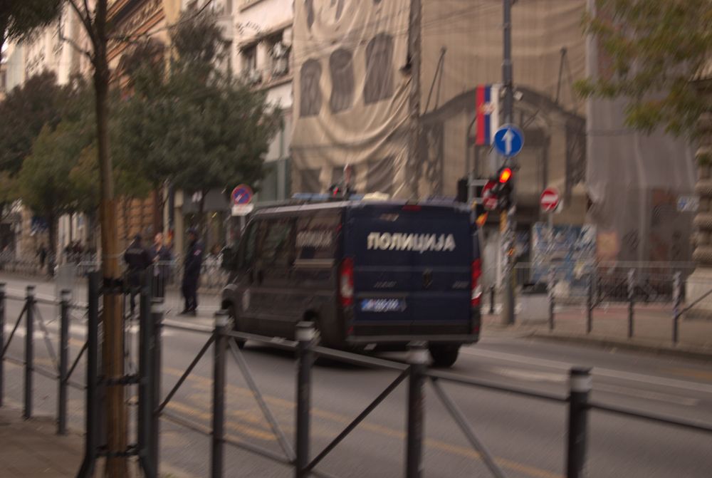 Tamni policijski kombi u Ulici kralja Milana u Beogradu, sa nekoliko policajaca u blizini, saobraćajnom signalizacijom i zgradom prekrivenom zaštitnom mrežom u pozadini.