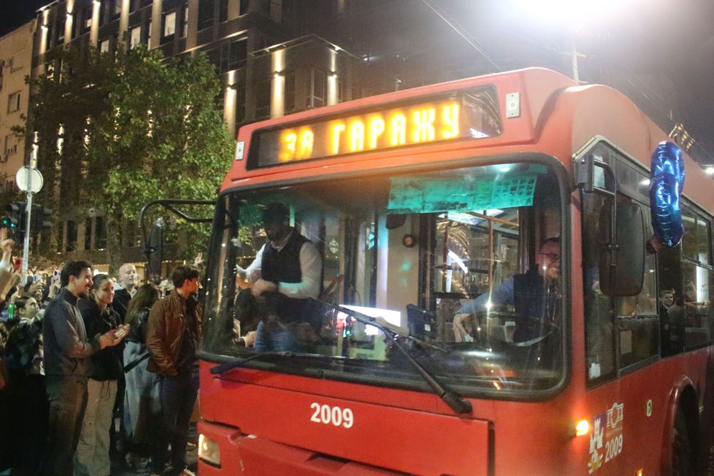 Crveni trolejbus sa oznakom „3A GARAŽA“ prolazi pored građana koji slave u Beogradu. Vozač se osmehuje, a pored prednjeg dela vozila vidi se plavi balon u obliku broja tri.