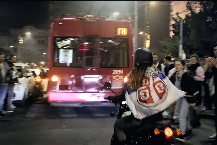 Devojka sa crnom kacigom na motociklu, ogrnuta zastavom Srbije, nalazi se iza crvenog trolejbusa 3A, dok građani stoje sa obe strane ulice u Beogradu tokom noći.