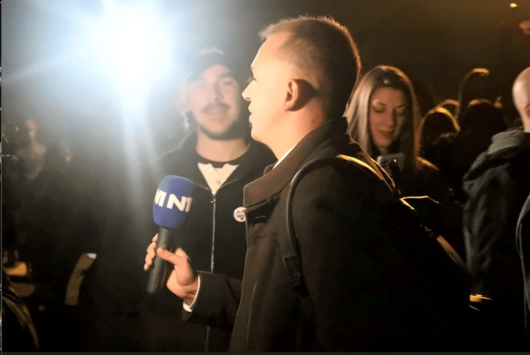 Reporter televizije N1 sa plavim mikrofonom razgovara sa mladićem u crnom kačketu u Pionirskom parku tokom noći, dok snažno svetlo osvetljava sagovornike, a iza njih stoje okupljeni građani.