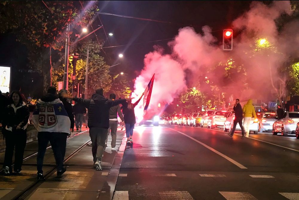 Grupa građana kreće se tramvajskim šinama u blizini Karađorđevog parka u Beogradu tokom noći. Jedan muškarac ogrnut je srpskom zastavom, drugi širi ruke, dok se ispred njih podiže crvenkasto osvetljen dim, a automobili stoje u koloni.
