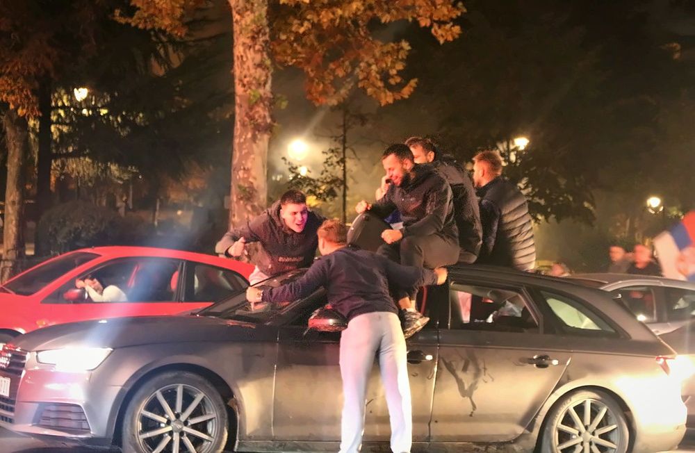 Grupa mladića okupljena oko sivog automobila na noćnoj ulici u Beogradu. Nekoliko njih sedi na vozilu ili se naginje preko njega, dok se levo vidi crveni automobil, a u pozadini drveće i gradska rasveta.