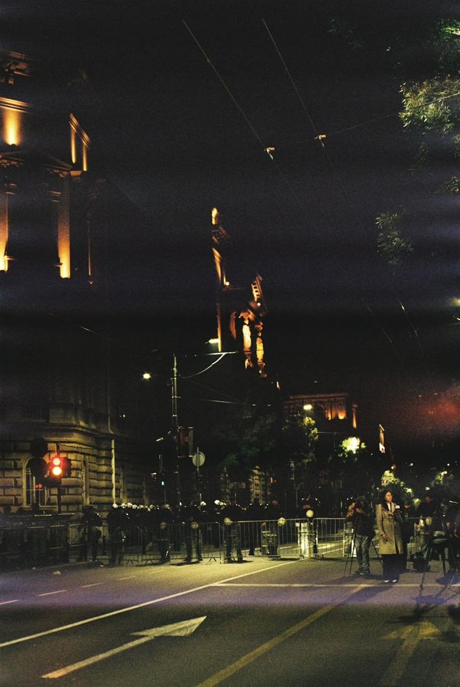 Police officers behind metal barriers on a street in central Belgrade at night, with illuminated buildings and a monument in the background.
