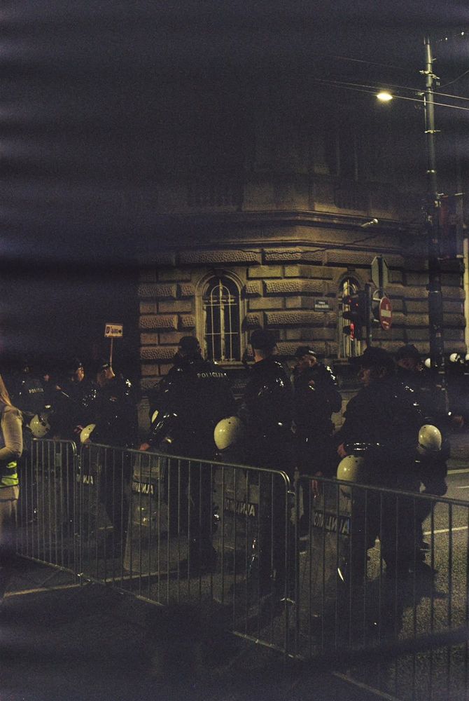 Police officers carrying protective shields stand behind metal barriers on a street in Belgrade at night.