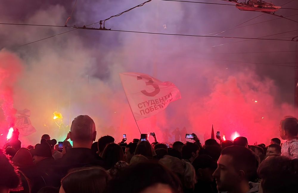 A crowd gathered at night amid red flare light and smoke, with a flag bearing the number three waving above them.