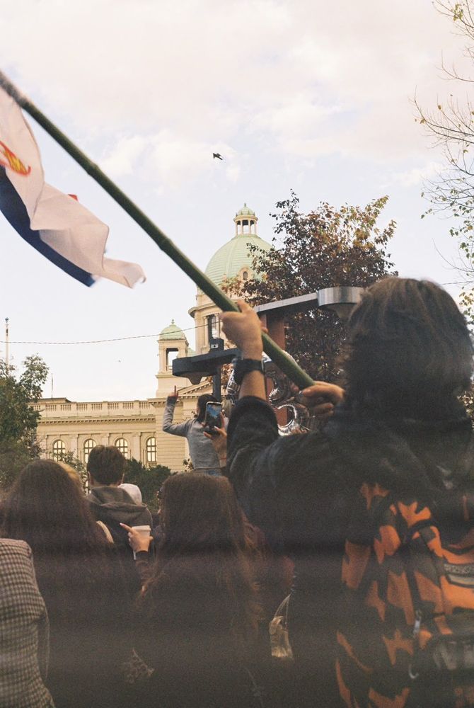 A person seen from behind raises a Serbian flag above a crowd, with the dome of the National Assembly building visible in the background in Belgrade.