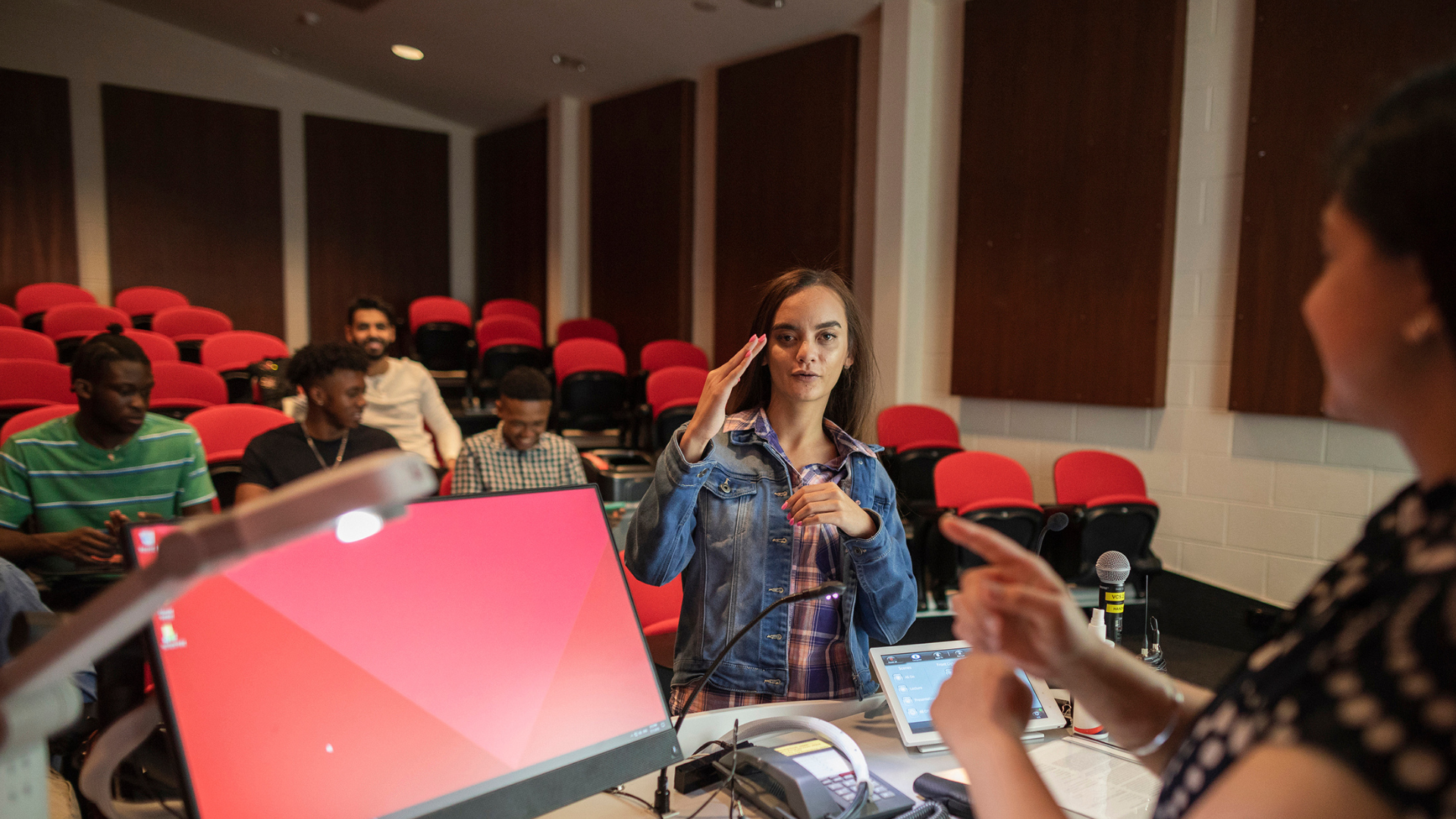 Young woman in lecture hall with a few other students sitting behind her as she uses sign language to communicate with the female professor, who is standing behind the lecture podium.