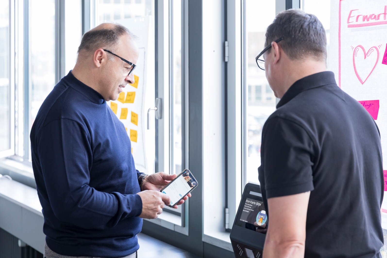 Zwei Kollegen im Gespräch über die Bedienung eines digitalen Terminals mit mobiler App