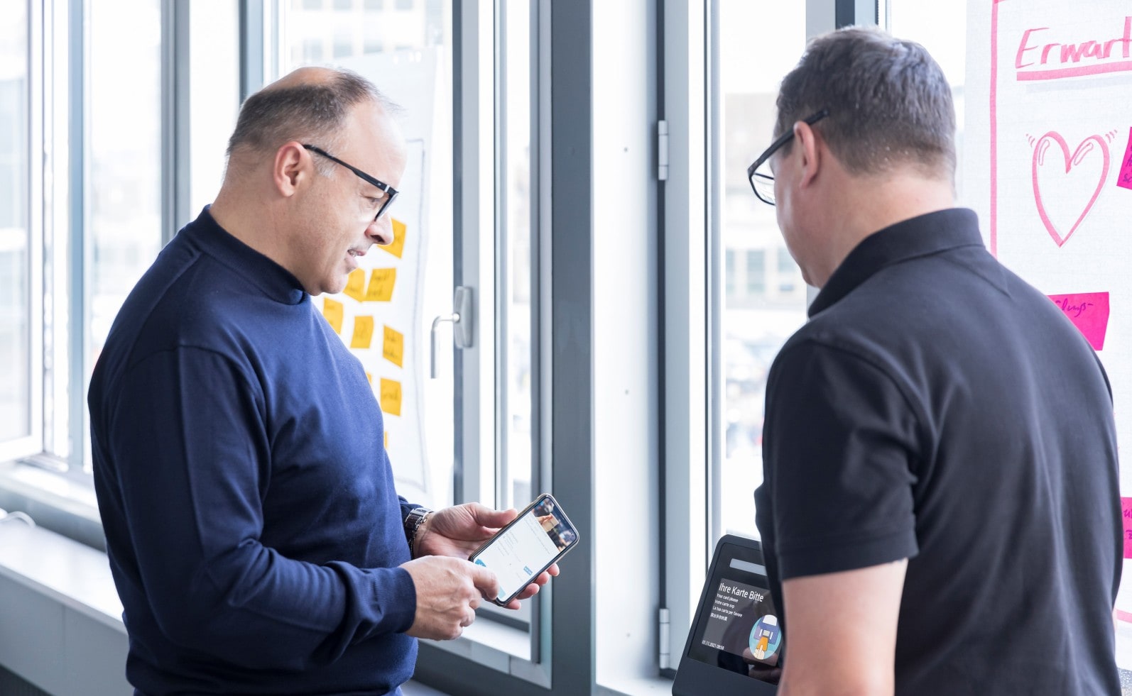 Zwei Kollegen im Gespräch über die Bedienung eines digitalen Terminals mit mobiler App