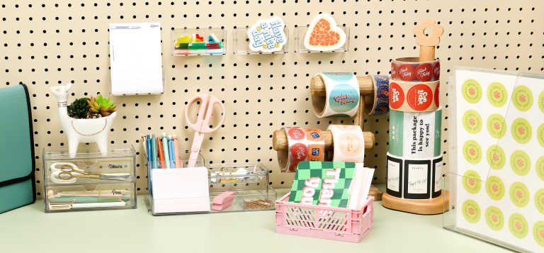 Organized desk workspace with a pegboard wall, clear acrylic organizers holding pens and supplies, labeled folders and drawers, and rolls of stickers stored neatly within reach