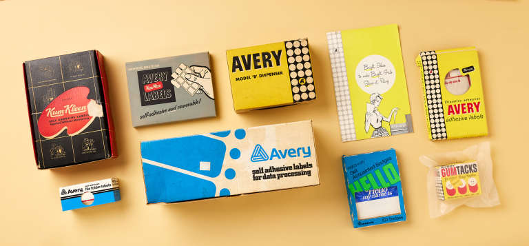 Flat lay of vintage Avery packaging from the mid-20th century arranged on a light tan background. The collection includes early adhesive label boxes, a label dispenser package, name badges, gum tack adhesive, and a marketing pamphlet, all featuring retro Avery logos and period graphic design. Some original sticker and label products remain inside the packaging, illustrating the early history of labels and stickers.