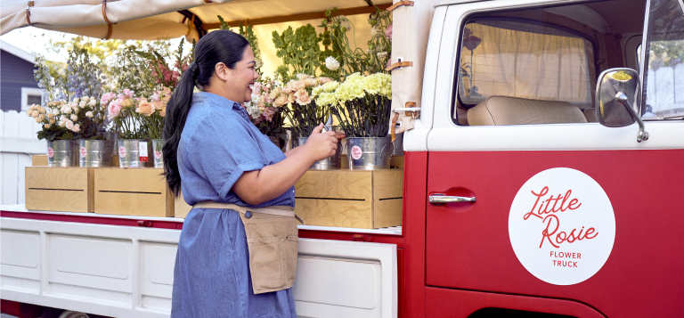 Owner Toni Rose beside her bright red Little Rosie Flower Truck with buckets of seasonal blooms displayed in the truck bed.