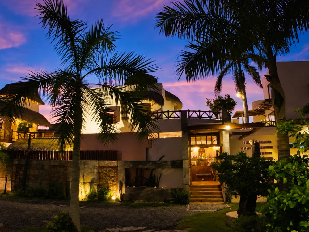 Fachada de A&V Hotel Boutique iluminada al anochecer, con techos de palapa entre palmeras