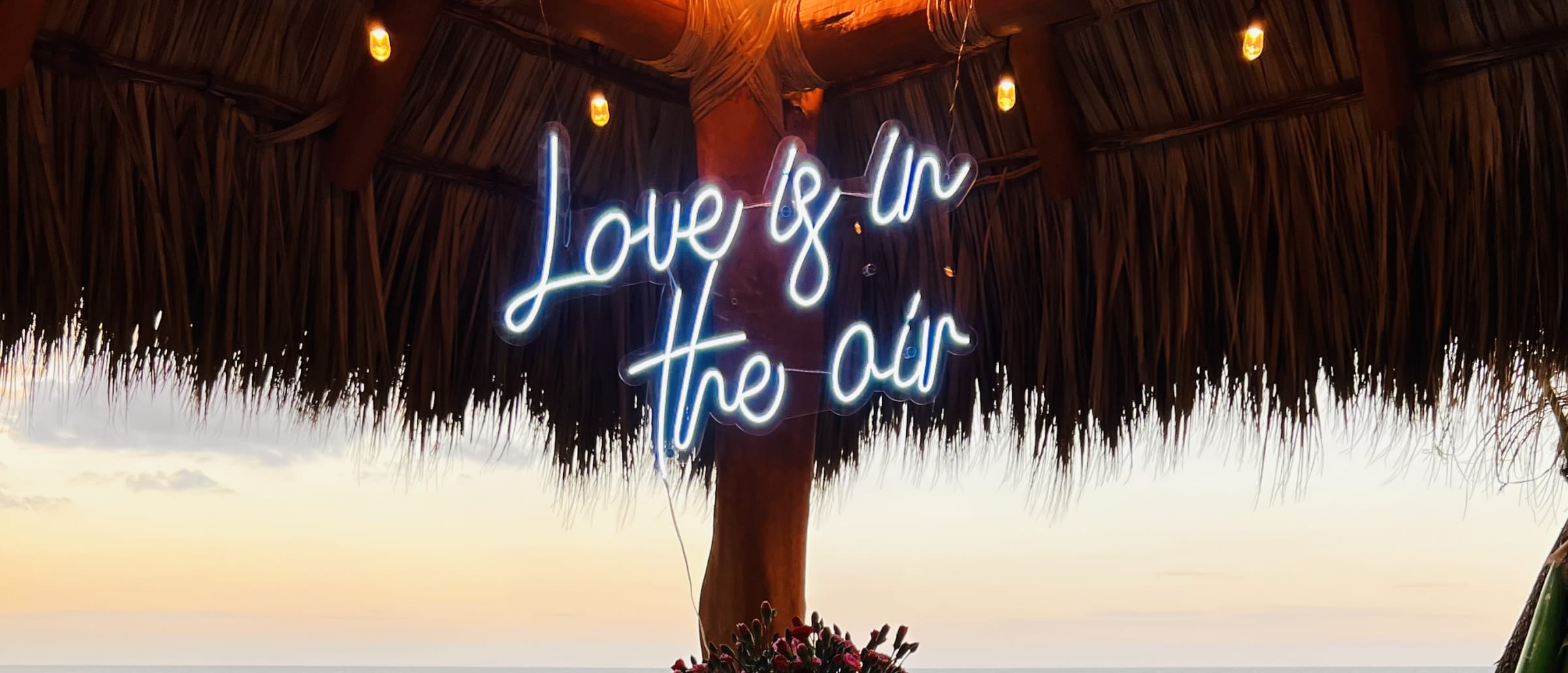 Mesa para dos bajo la palapa frente al mar al atardecer, con el letrero «Love is in the air»